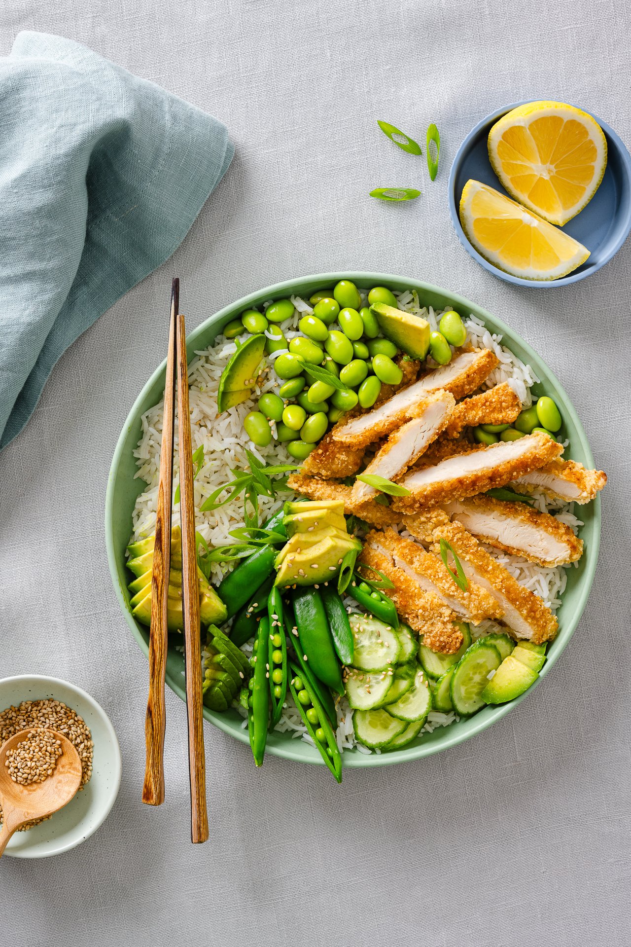 an asian inspired bowl with crispy chicken tenders, rice, edamame, snap peas, pickled cucumber, avocado and green onions with chopsticks resting on top and a little bowl of lemons and sesame seeds beside it