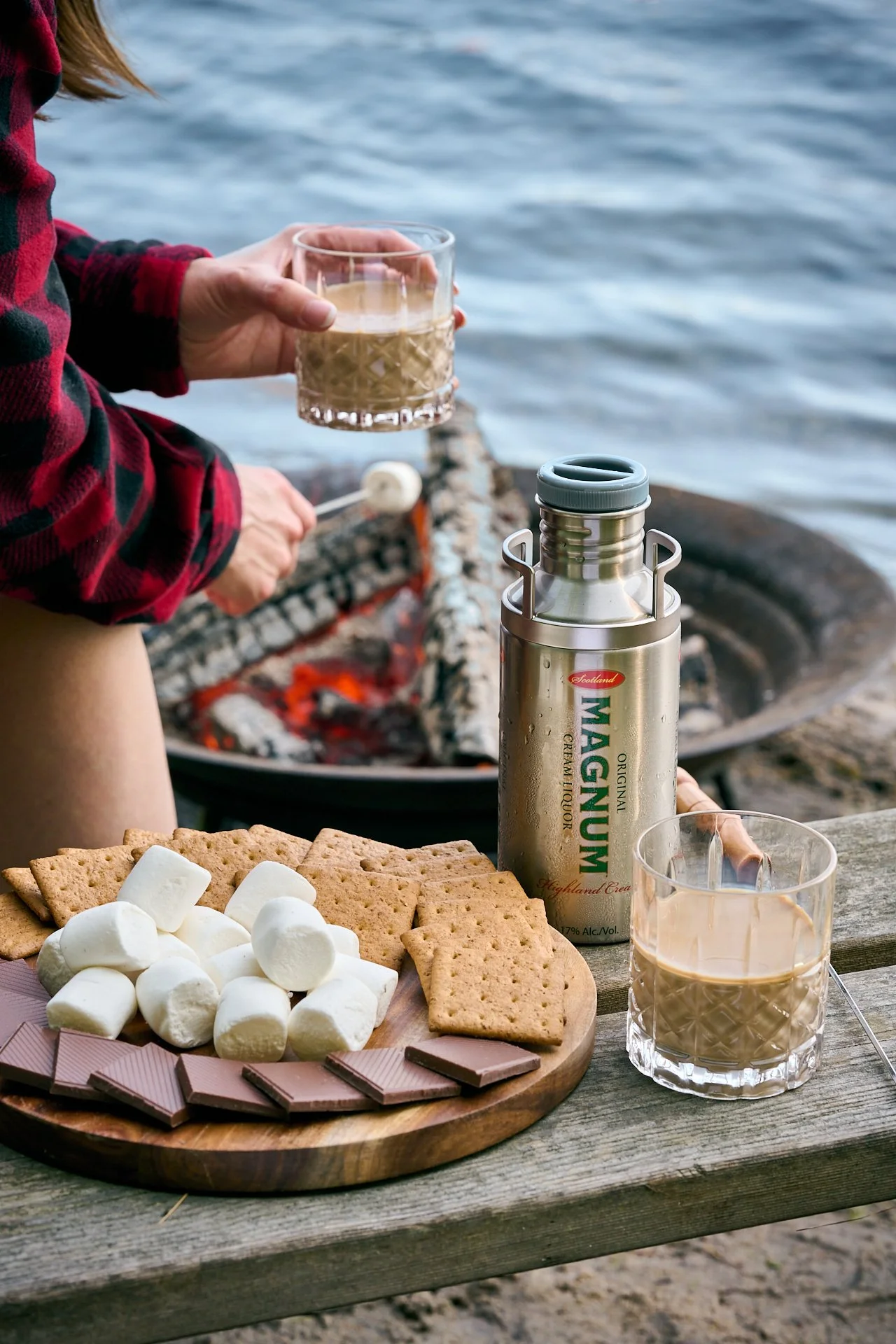 a person resting their hand on a knee holding a glass of magnum cream liqueur, beside them is a fire and the edge of a lake with a platter of s'more ingredients like graham crackers, marshmallows and chocolate resting on a plate