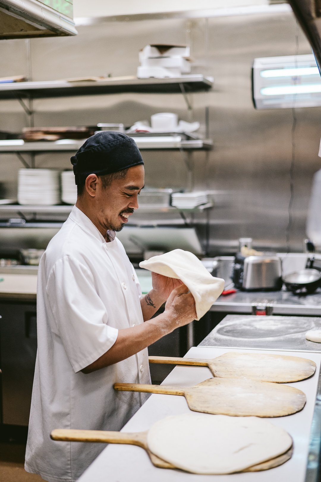 chef at a pizza station in an italian restaurant, he's wearing a white chef's coat and a black chef's hat, he's smiling as he's shaping the pizza dough into a circle