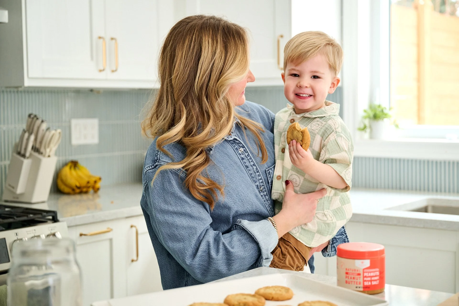 mom holding her toddler with homemade peanut butter cookies and nate's peanut butter in a kitchen