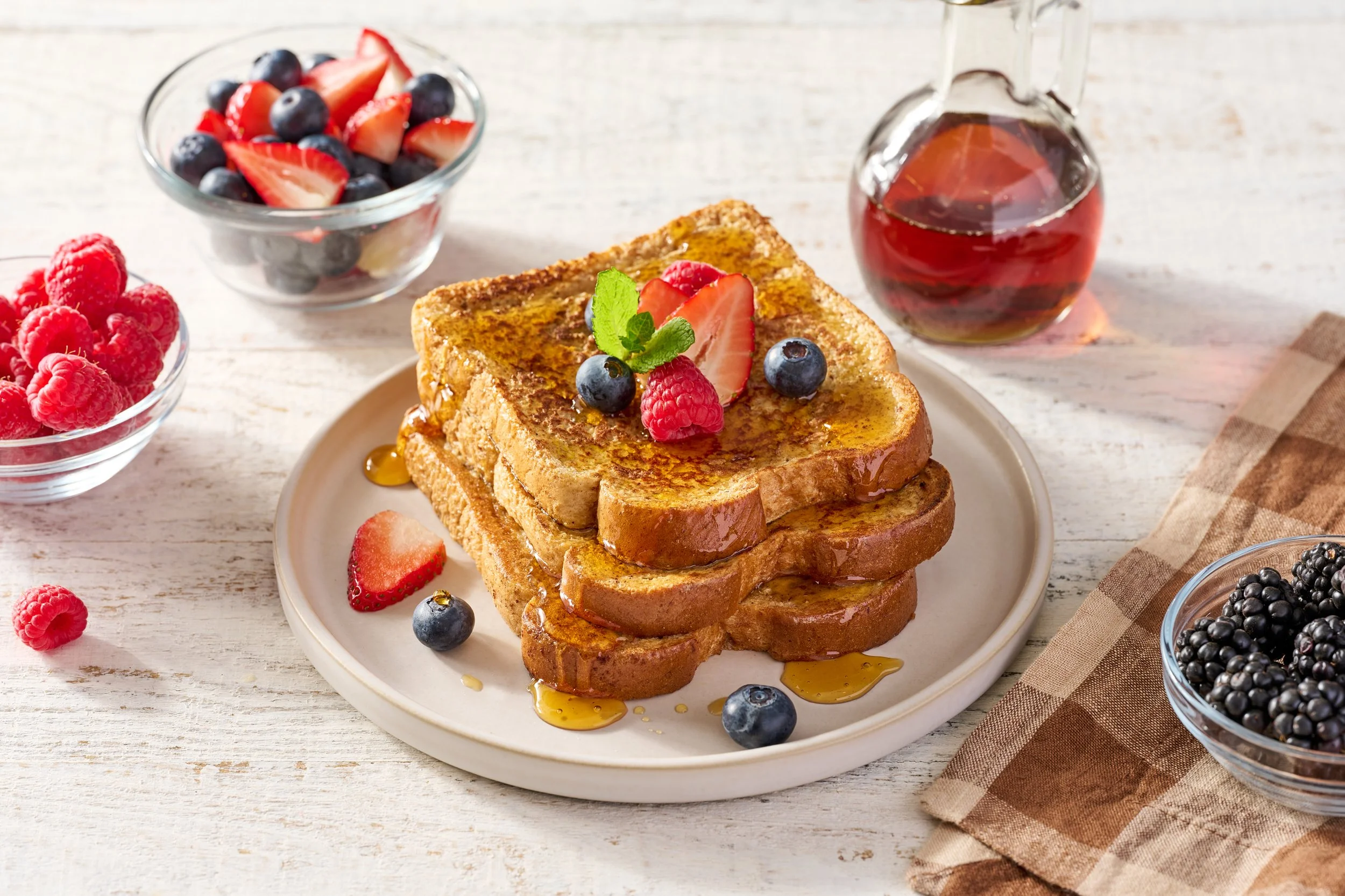 a stack of french toast on a plate with maple syrup dripping down with bows of fresh berries beside it on a rustic wood surface