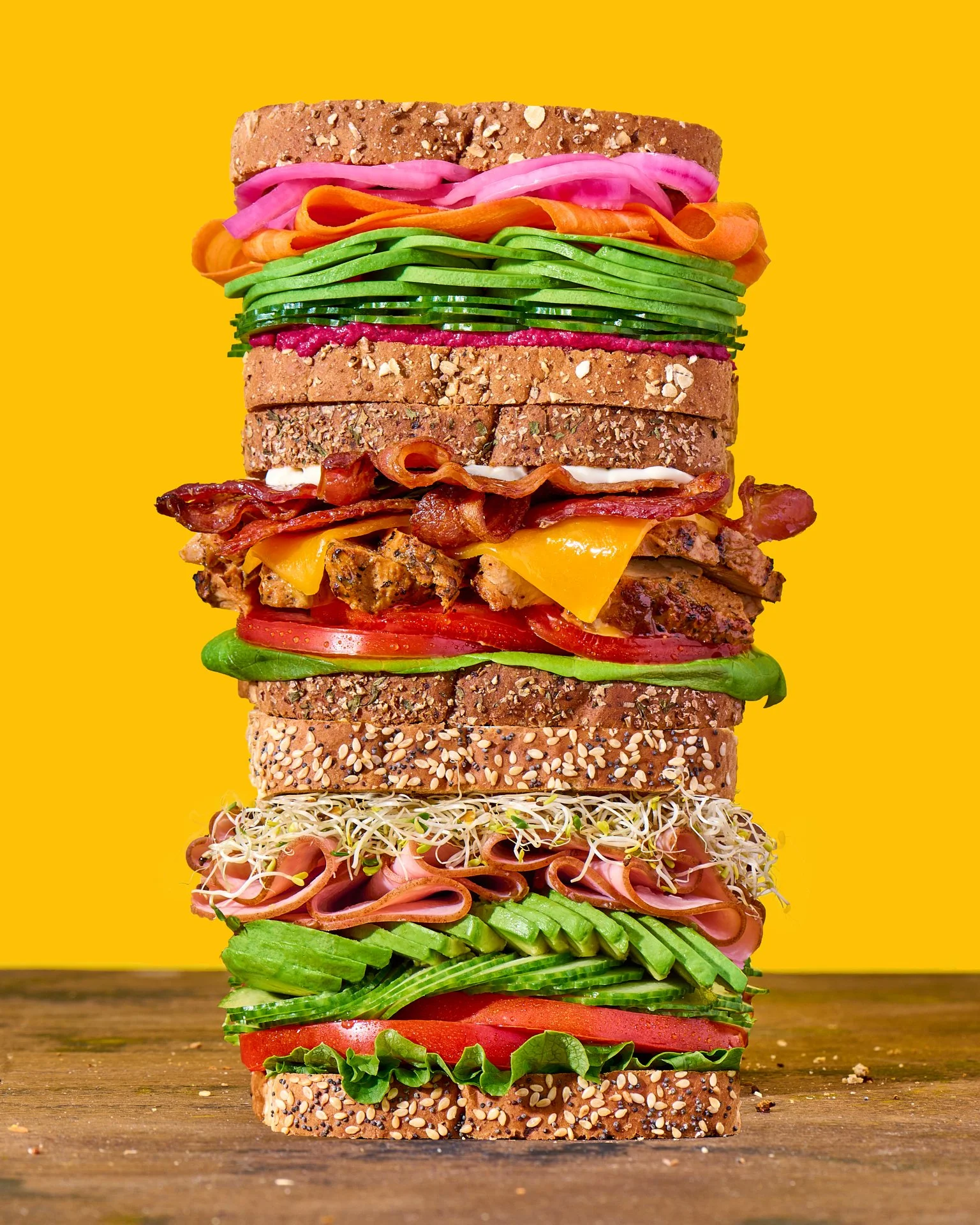 a towering sandwiches stack with 3 sandwiches, layers of meat and cheese, avocado and pickled radishes, and sliced meat and sprouts on a wooden surface and orange background