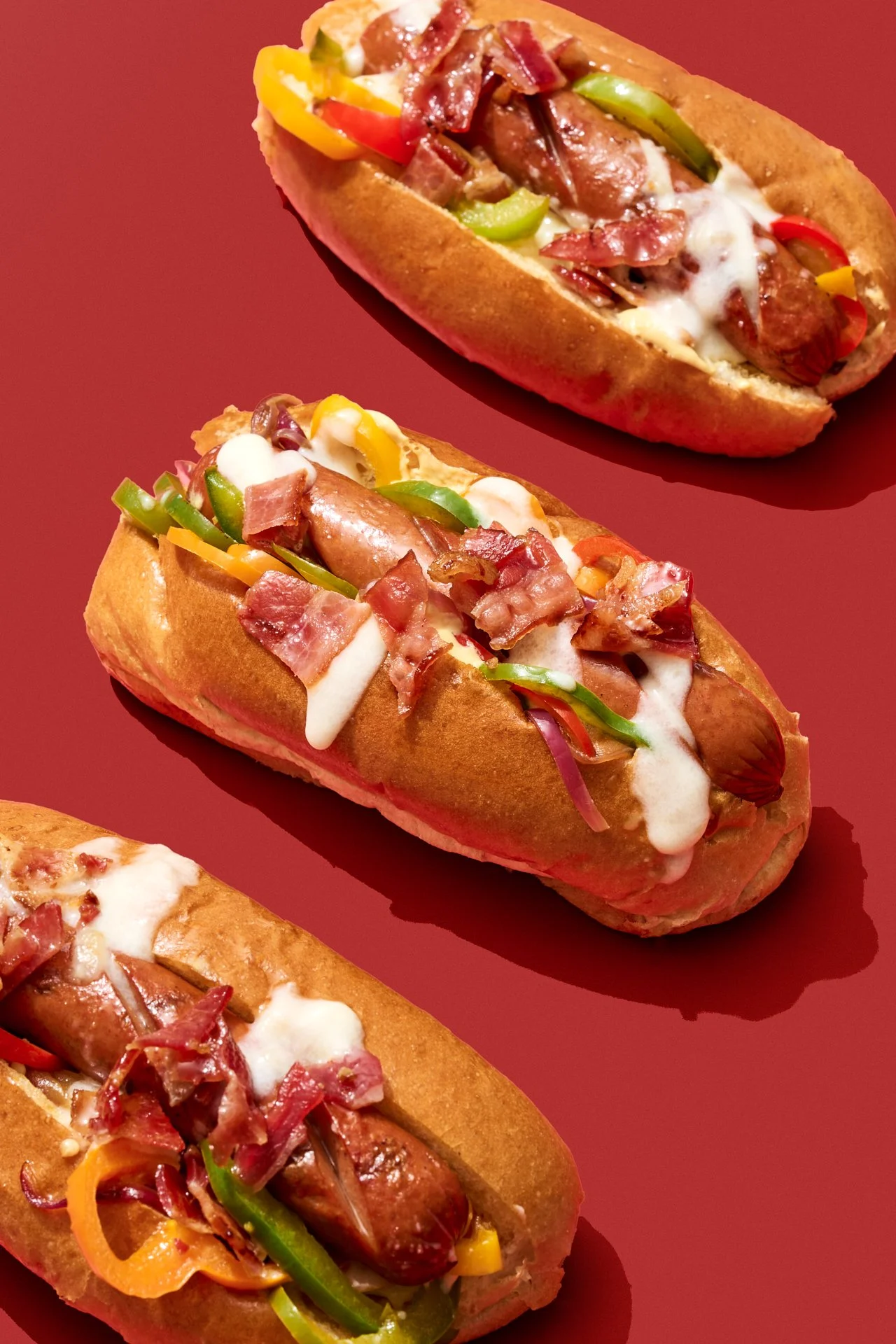 three maple leaf hot dogs in a bun with sauteed peppers, bacon, and a drizzle of white sauce, commercially shot on a red backdrop