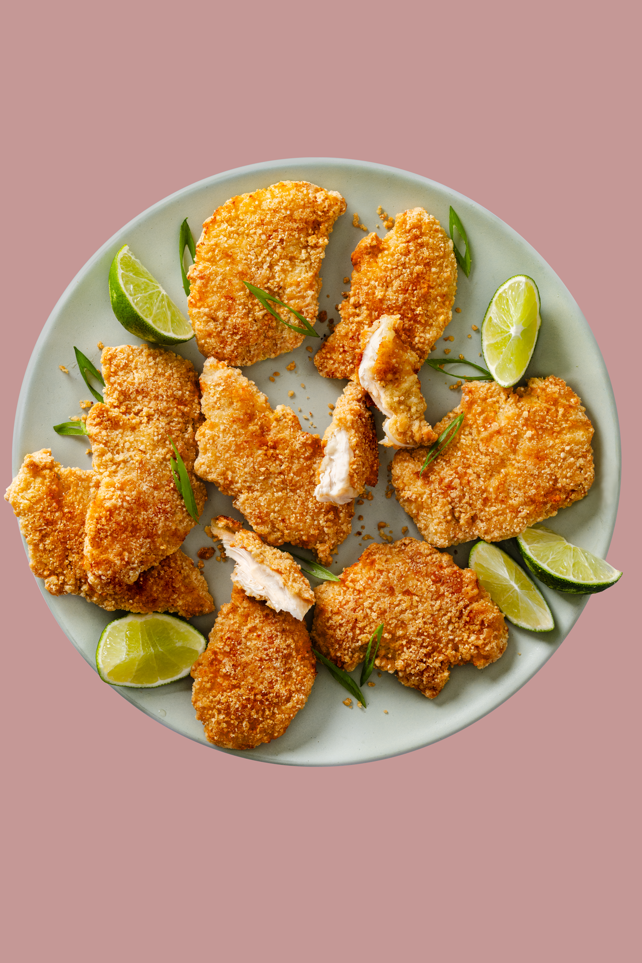 Fried chicken pieces garnished with lime wedges and green onion slices on a pale green plate with a pink background.