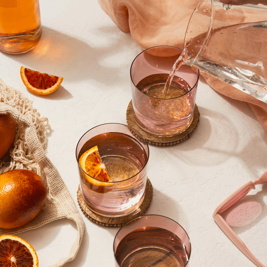 a bright sunny stopm otion with kilne's pink glassware surrounded with blood oranges and pink sunglasses. a pitcher of water is pouring into one of the glasses
