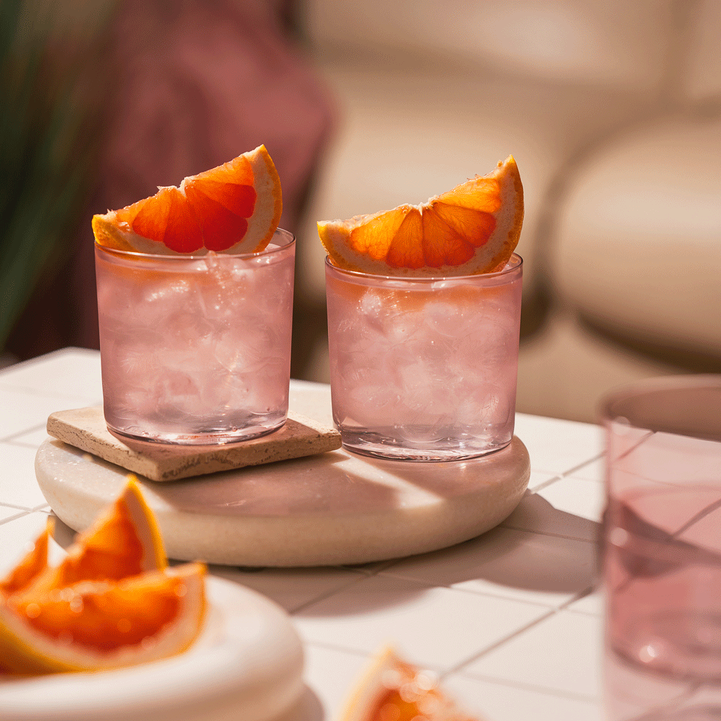 a stop motion of pink glasses with pink grapefruit resting on top and sitting on a tile surface with a hand coming in and taking one out