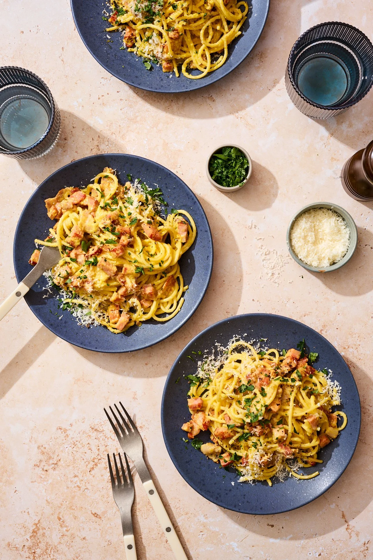 dark blue plates with carbonara and bacon on top with a sprinkle of parmesan and chopped herbs. beside them is a glass of water, utensils, a bowl of parm and some fresh herbs on a warm stone backdrop