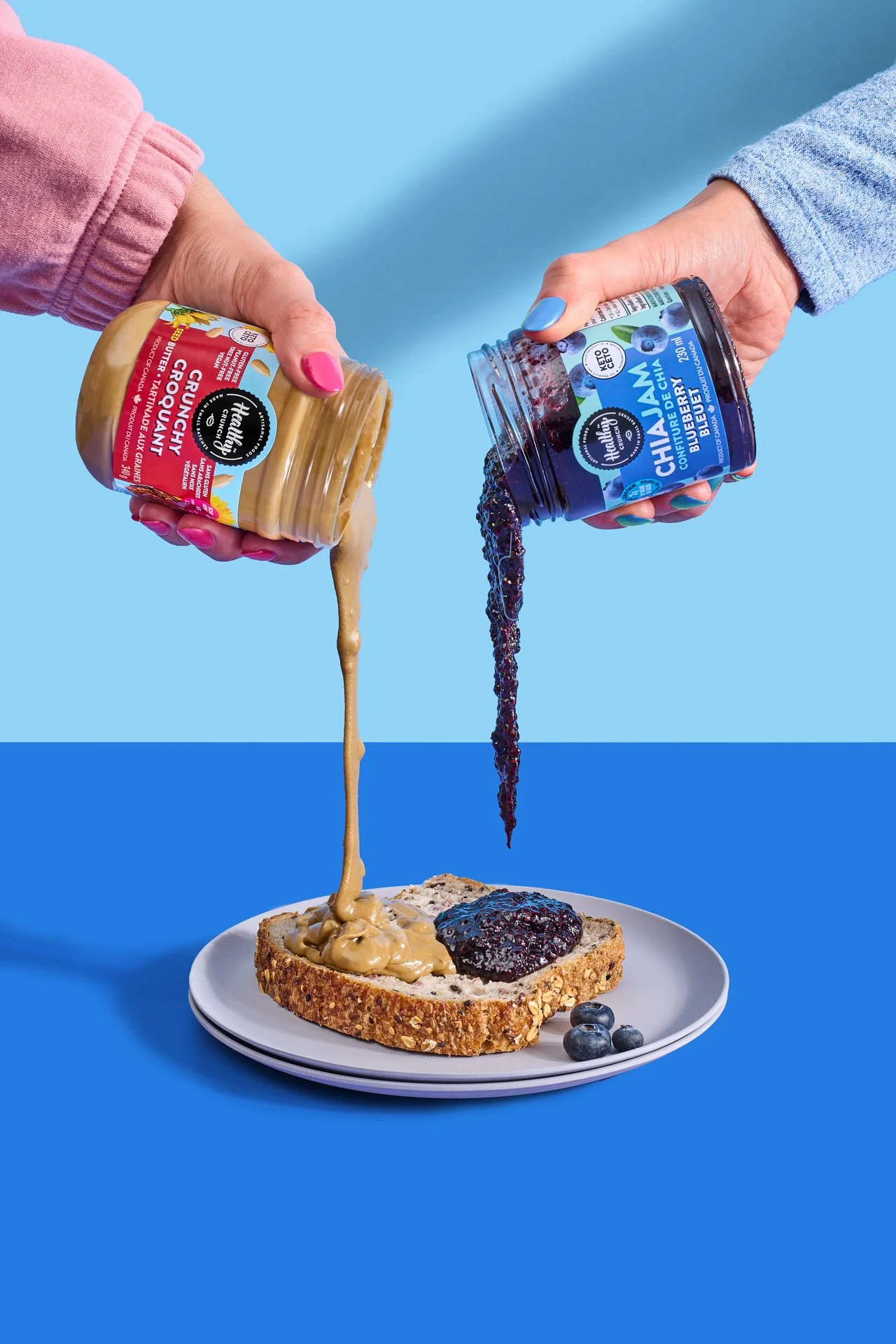 two hands pouring nut butter and jam on top a piece of toast to make a pb & j sandwich on a blue surface