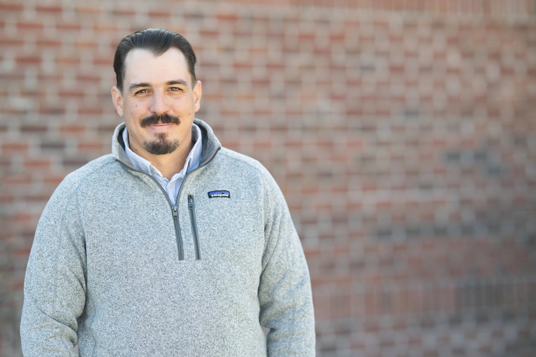 Portrait of a man with dark hair, mustache, and goatee, wearing a gray Patagonia fleece jacket standing outdoors against a brick wall.