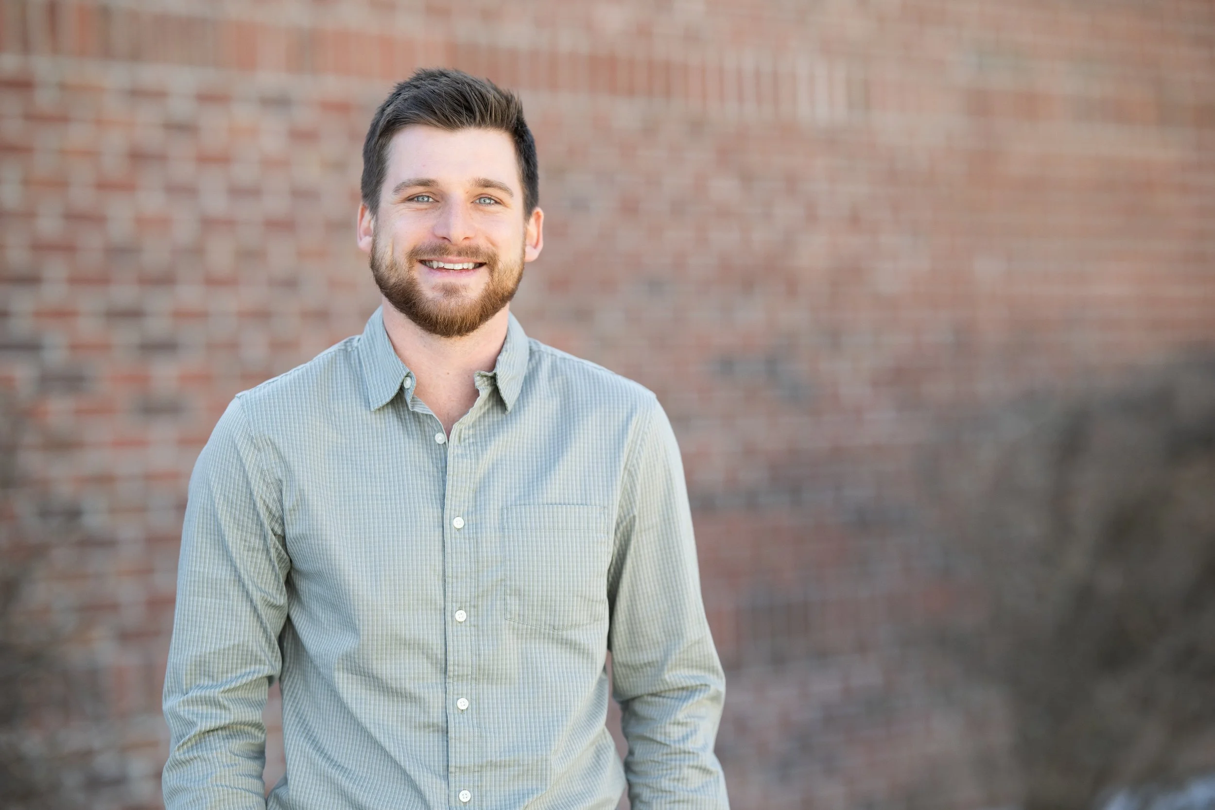 A smiling man with dark hair and a beard posing outdoors in front of a brick wall.