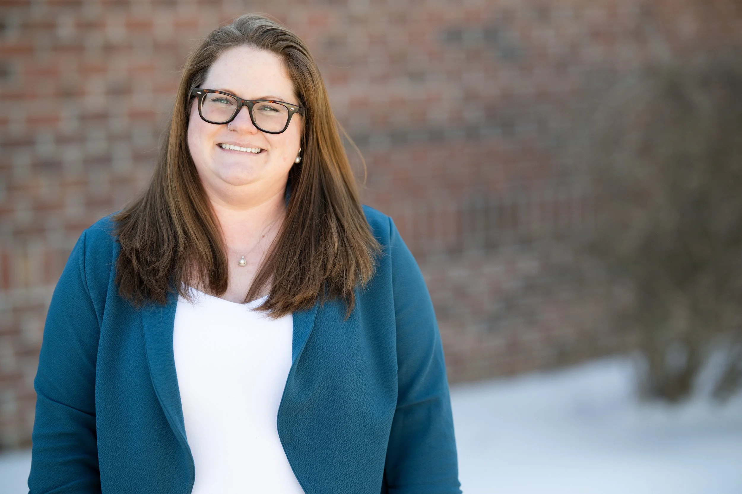 A woman with brown hair, glasses, and earrings smiling outdoors in front of a brick wall and snow.