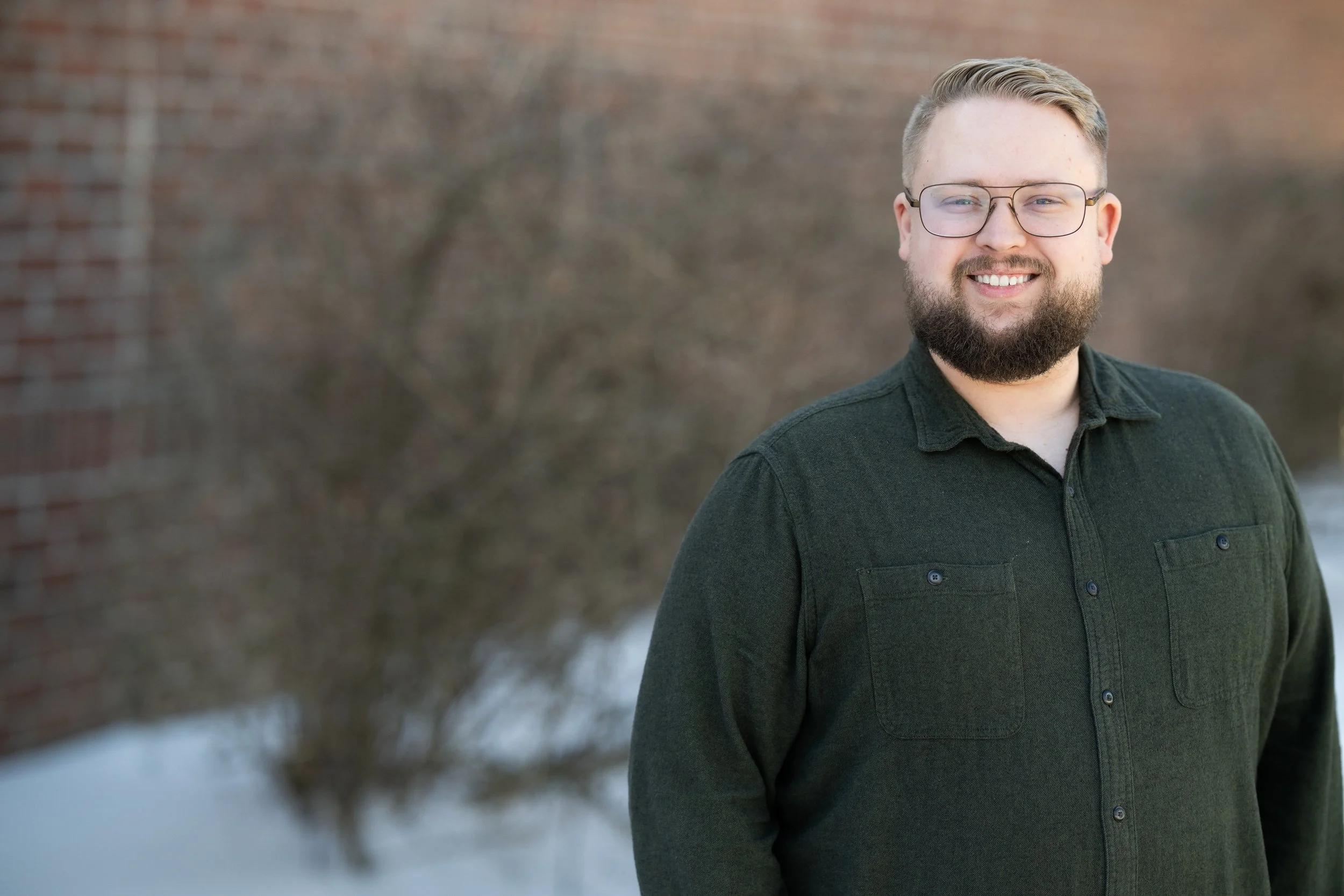 A smiling man with glasses and a beard standing outdoors in winter with snow on the ground and a blurred brick wall and tree in the background.