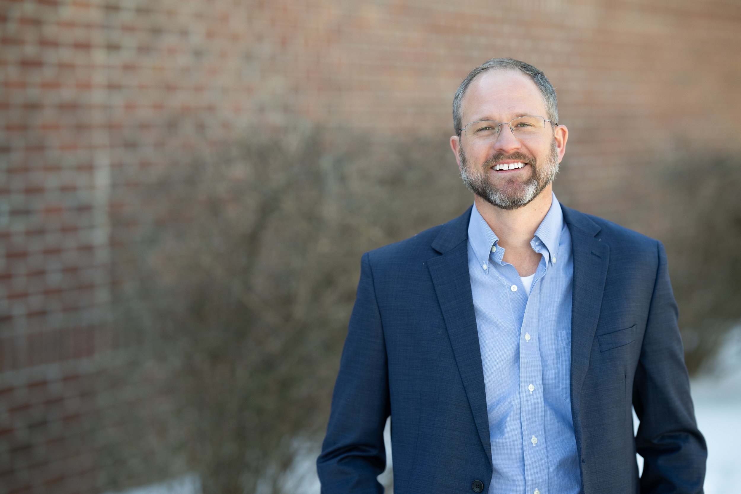 A smiling man with glasses, a beard, dressed in a blue blazer and light blue shirt, standing outdoors near a brick wall and some bare bushes.