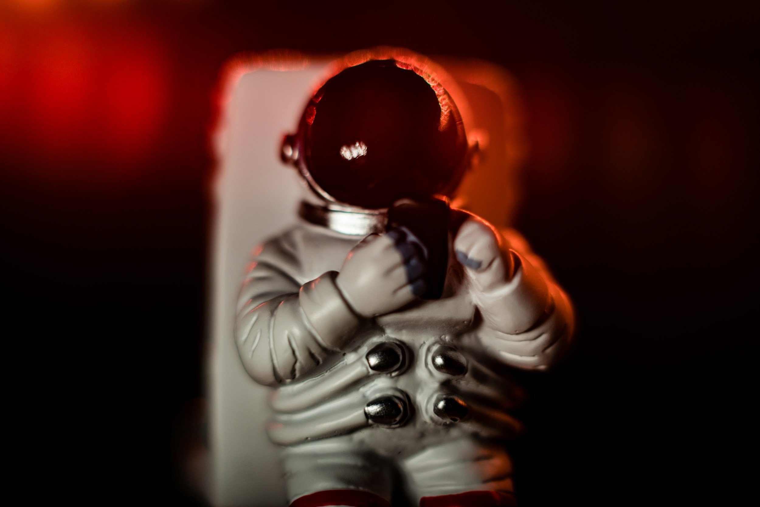 Close-up of a toy astronaut holding a small black camera, with a dark background and red lighting.