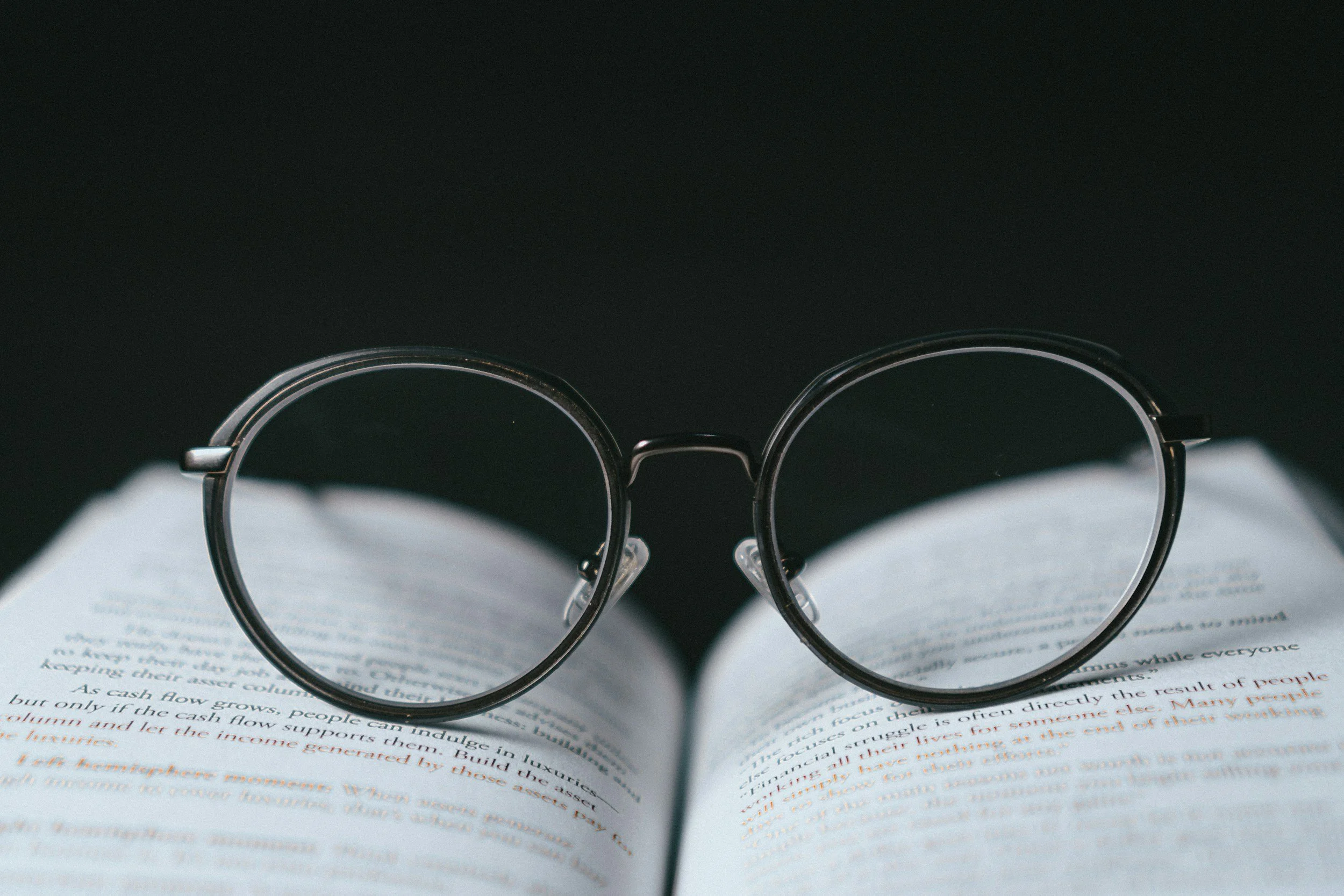 A pair of round eyeglasses resting on an open book with black background.