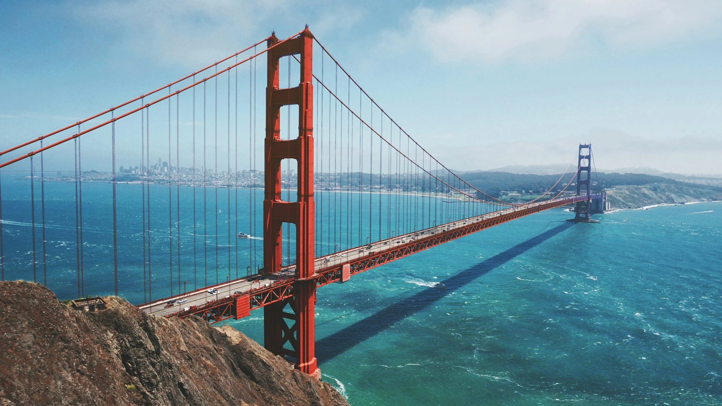 Aerial view of the Golden Gate Bridge in San Francisco, California, with cars on the roadway and the city skyline in the background, turquoise water below, and a blue sky.