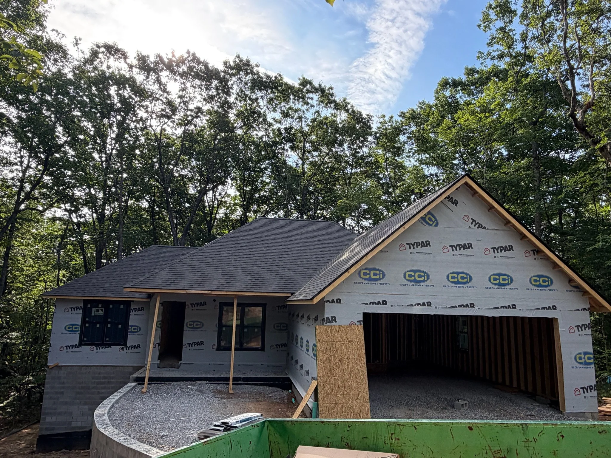 A house under construction with a partially finished exterior, including windows and a roof, surrounded by trees and a clear sky.