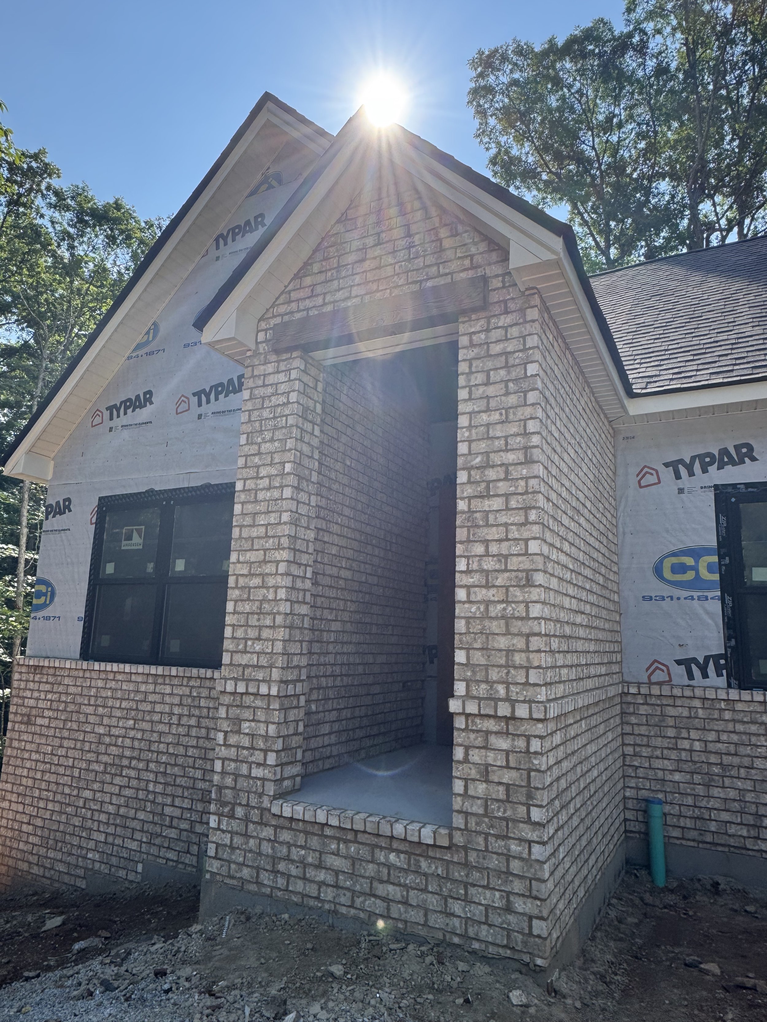A house under construction with brick exterior walls, a covered patio, and a steep gable roof. The house is partially wrapped in building materials, with visible windows and a small porch. The sun is shining brightly overhead, creating lens flare.