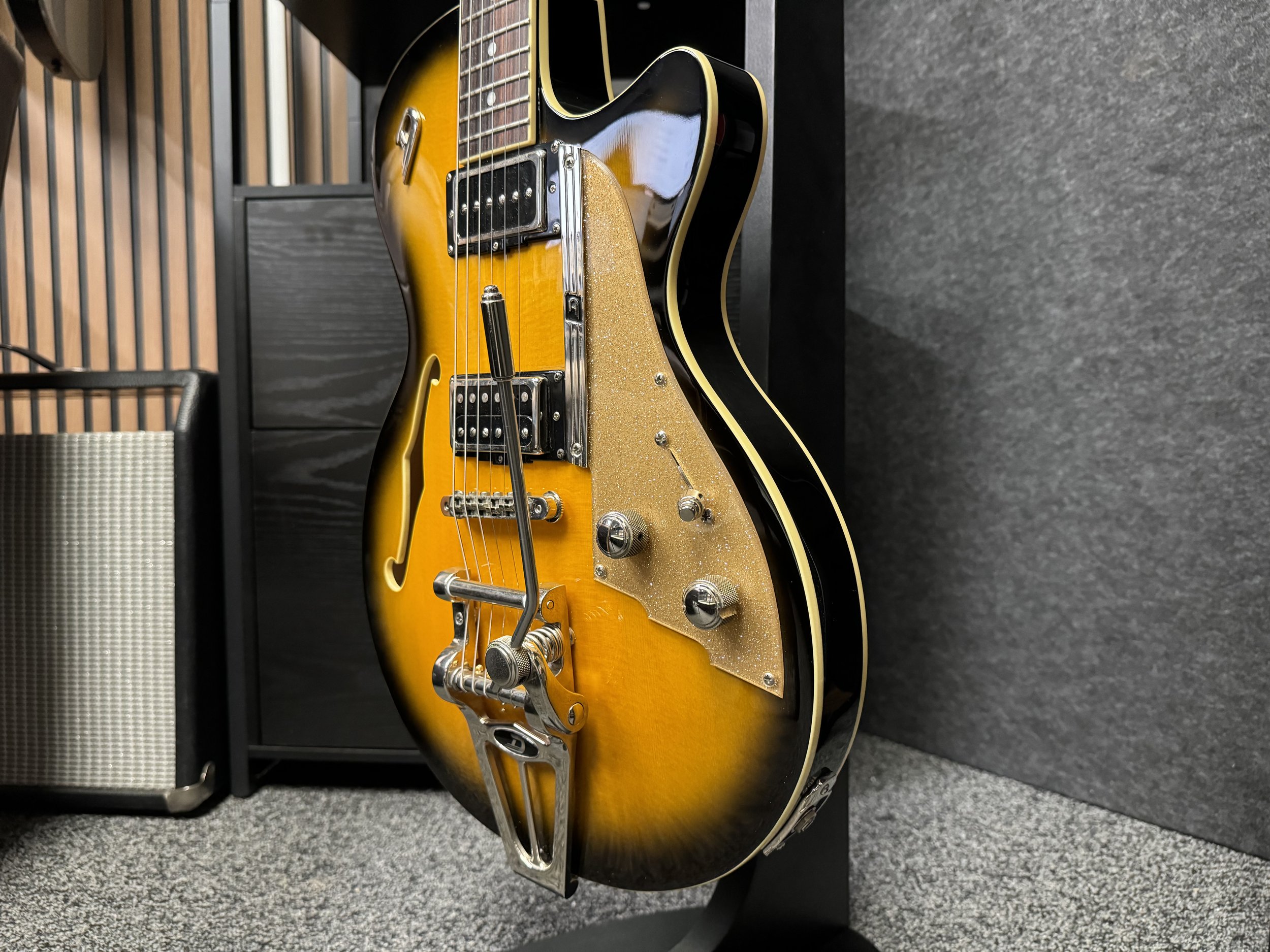 Sunburst semi-hollow electric guitar on a stand in a room, with an amplifier in the background.