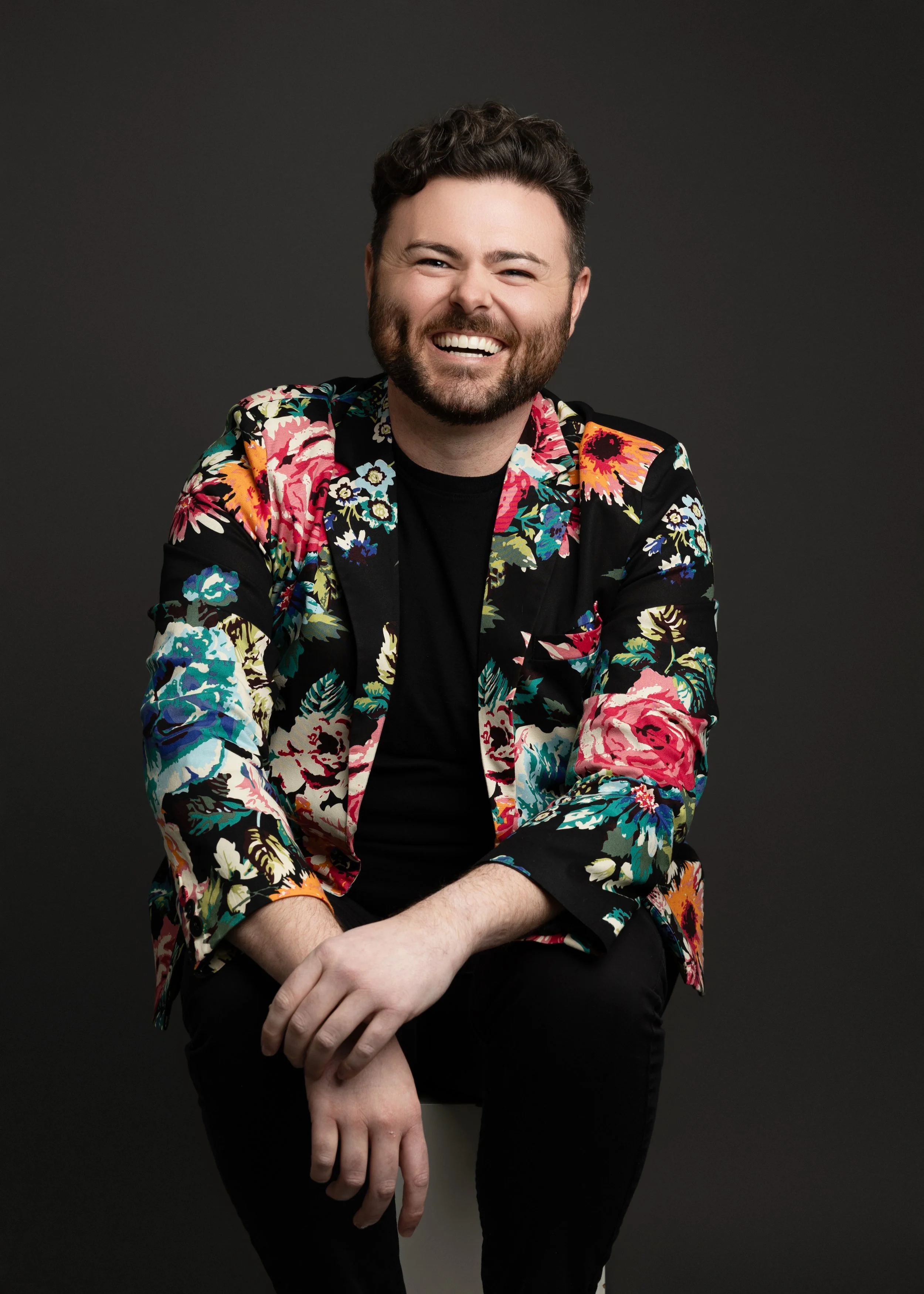 A smiling man with dark curly hair and a beard, wearing a colorful floral blazer over a black shirt, sitting against a dark background.