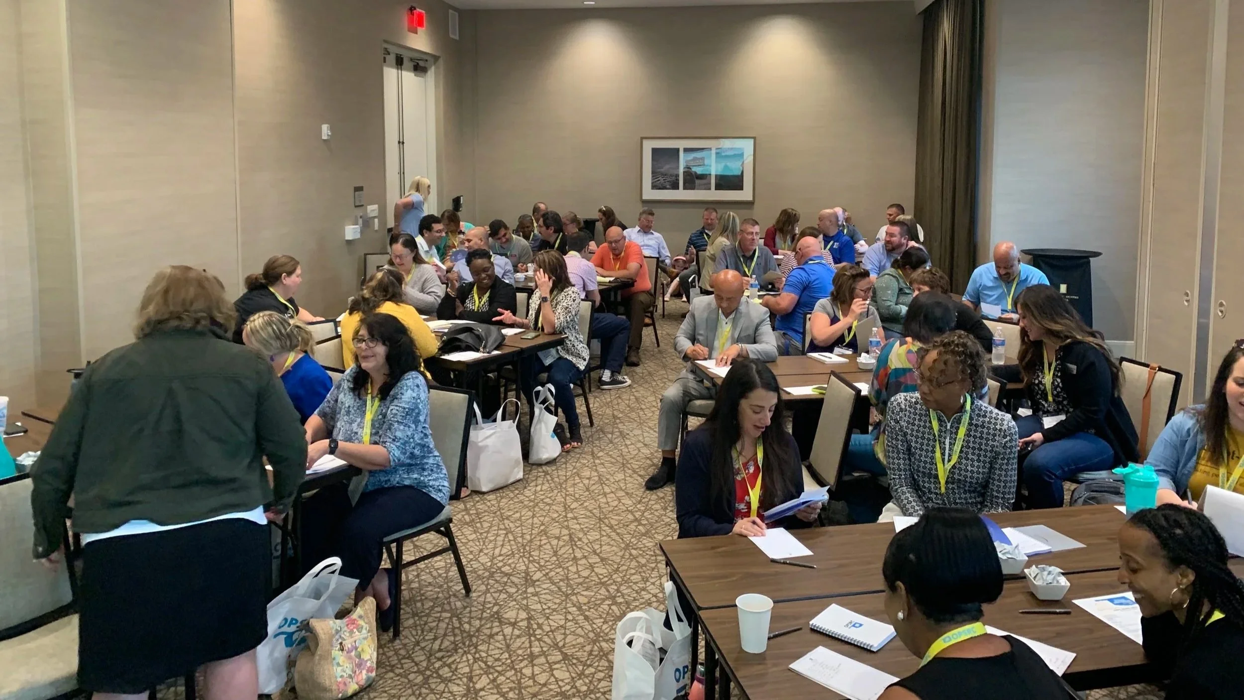Conference room filled with diverse attendees seated at tables, engaging in discussions and reviewing documents during a professional event.