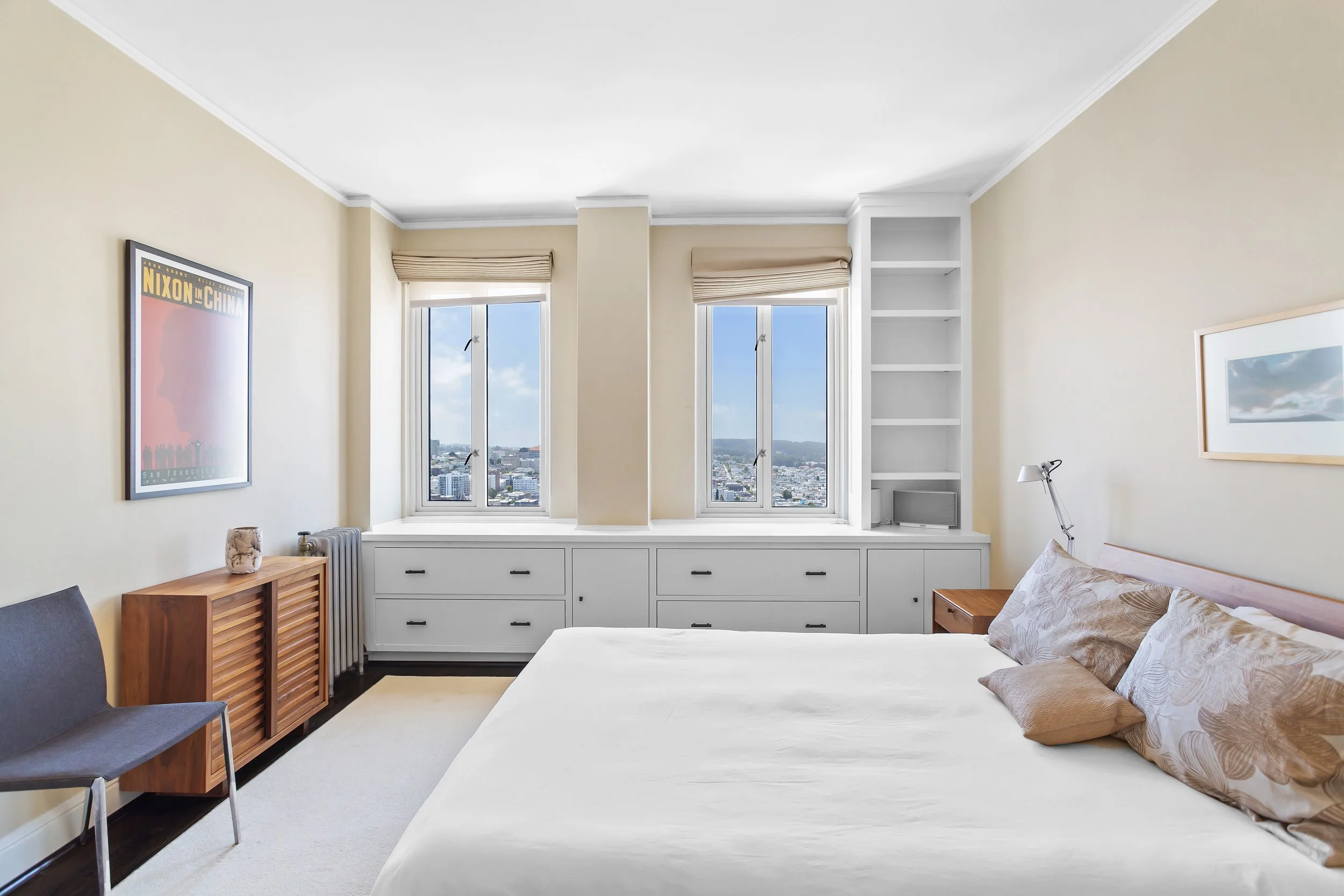 Modern bedroom with beige walls, large windows with city view, white built-in storage, bed with floral pillows, artist print on wall, side table, and small armchair.