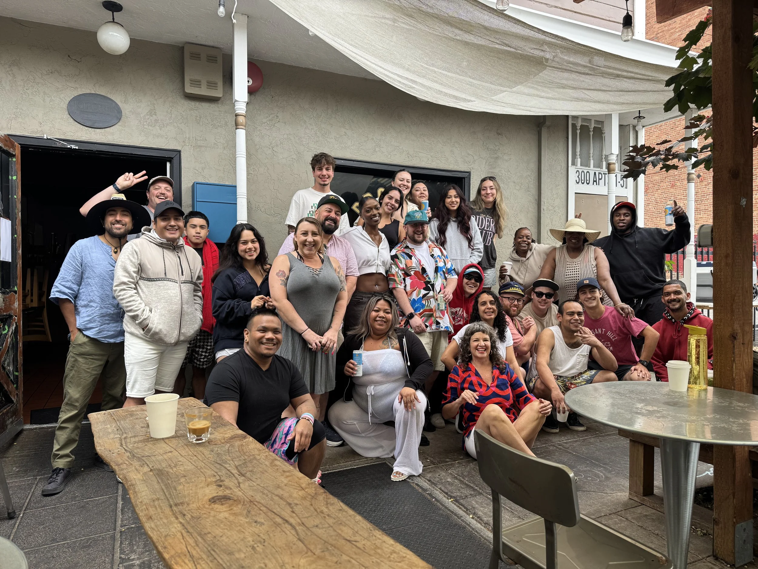 A group of around 20 diverse people gathered outdoors on a patio, smiling and posing for a group photo. Some are holding drinks, and they appear to be celebrating or at a social gathering.