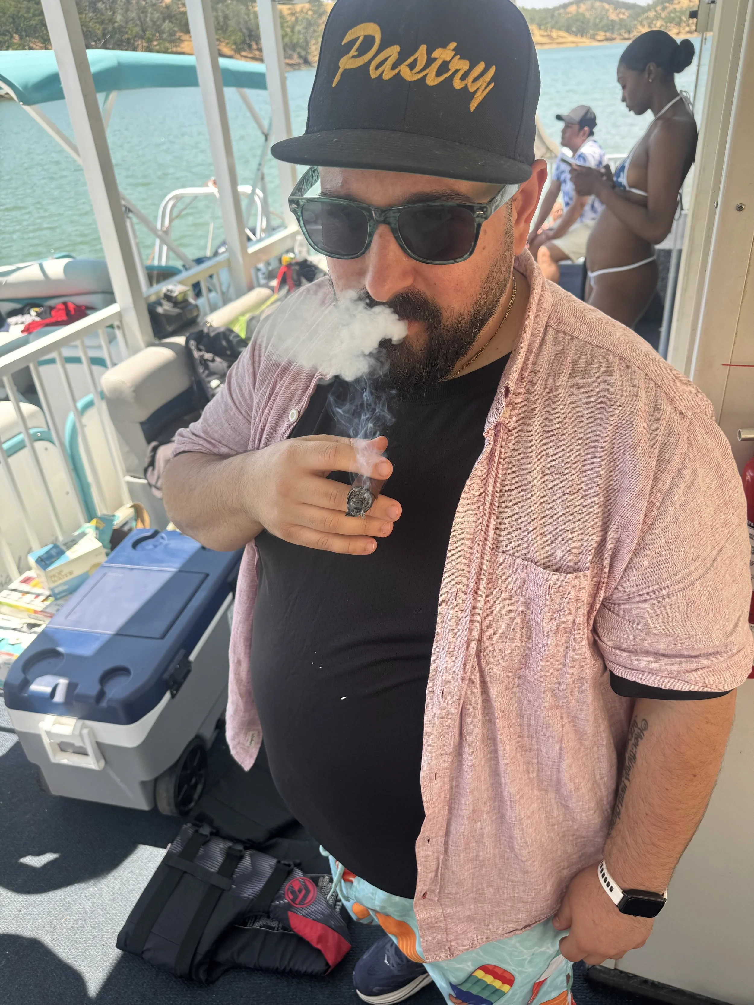 Man wearing sunglasses and a hat that says 'Pastry', smoking a cigar on a boat with water in the background, and other people on the boat.