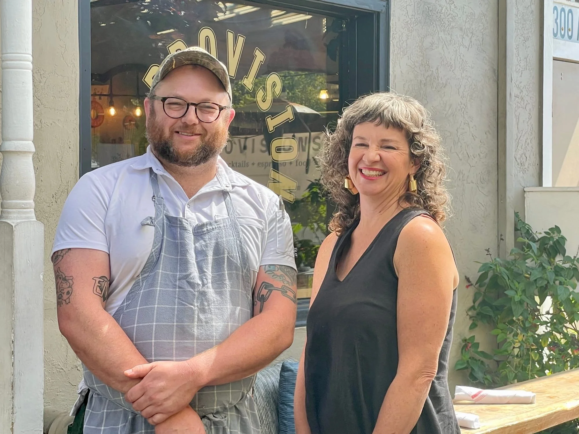 A man with glasses, a beard, wearing a white shirt and gray apron, standing next to a woman with curly hair, wearing a black sleeveless top, both smiling outdoors in front of a restaurant window.