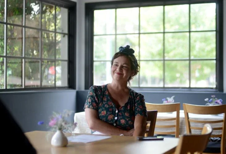 Woman with a floral dress and hair tied up with a bow, smiling and sitting at a table in a sunlit room with large windows and wooden chairs.