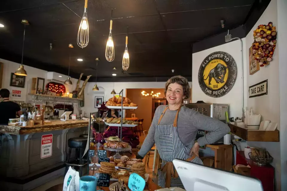 A smiling woman behind a counter in a coffee shop or bakery, wearing an apron with blue and white stripes, surrounded by baked goods like cookies and pastries, with a barista in the background, and various signs and decorations on the wall.