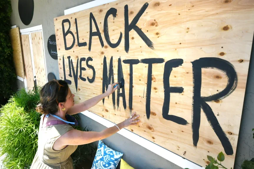 Woman painting the words 'Black Lives Matter' on a wooden sign outdoors.