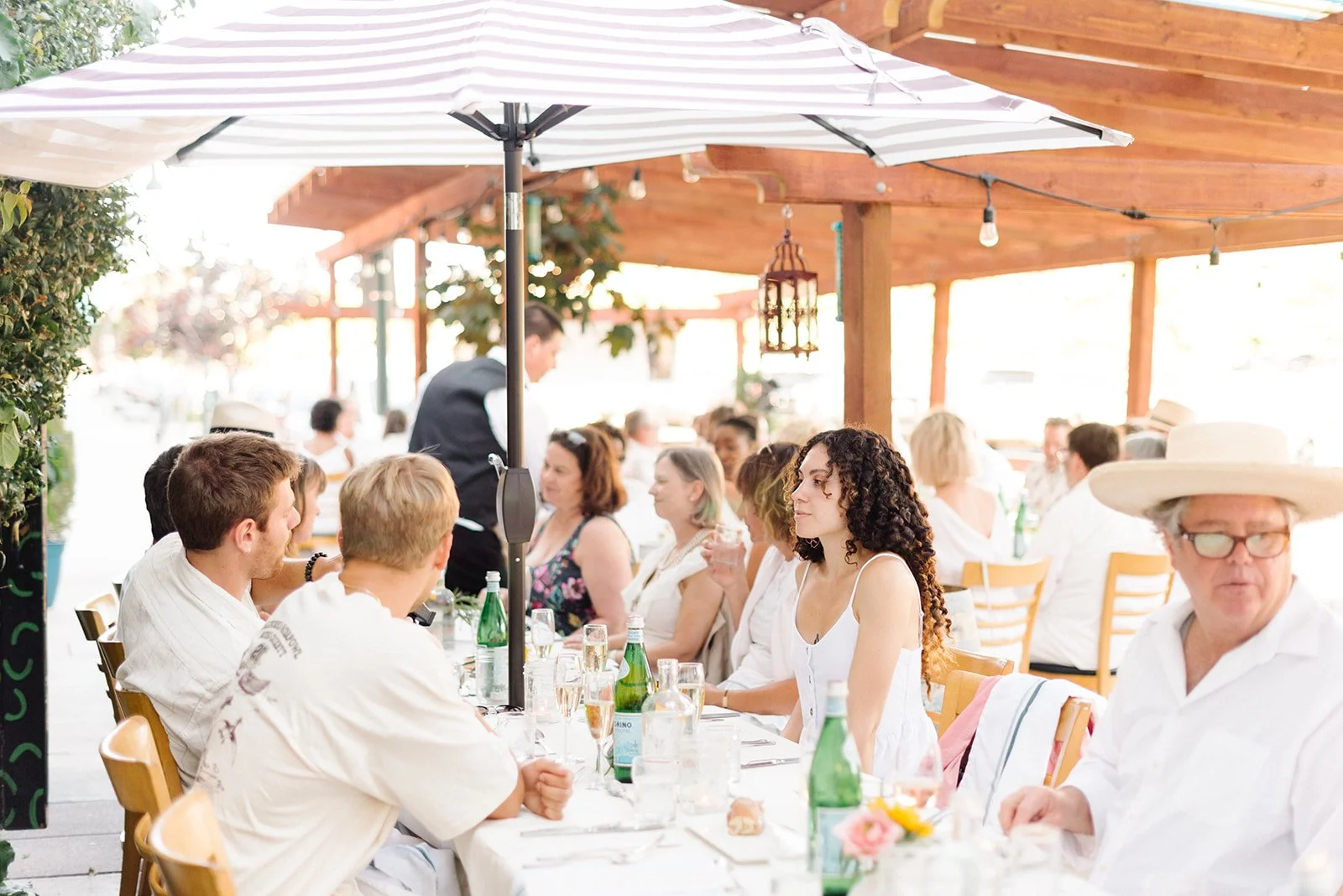People dining outdoors under a large umbrella, with a long table set with drinks and decorations, at a casual outdoor restaurant or gathering.