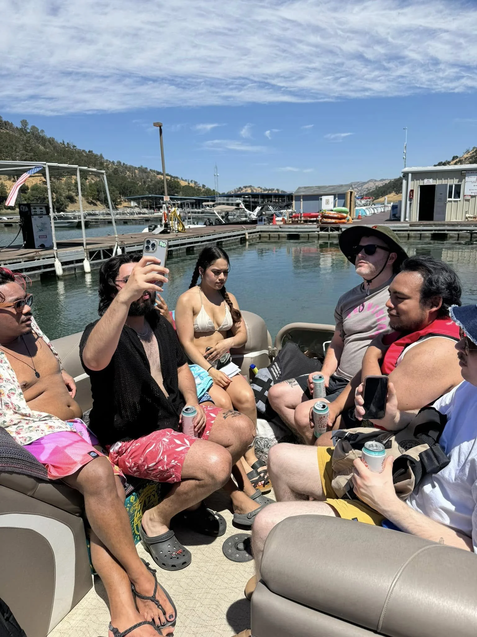 Group of people sitting on a boat near a marina, some holding cans, taking photos, dressed in swimwear and casual summer clothing, with boats and mountains in the background.