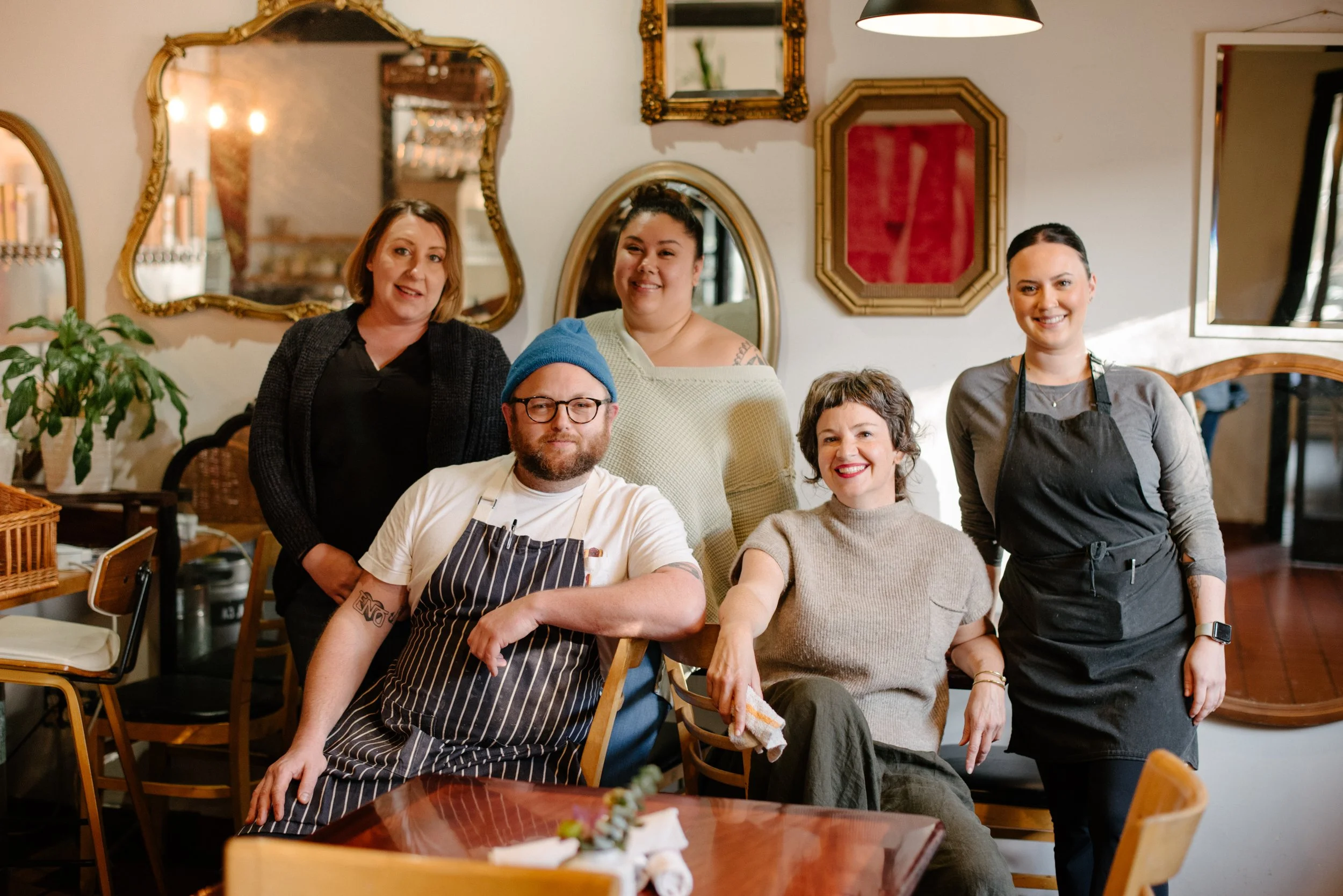 Group of five restaurant staff members, including chefs and waitstaff, smiling inside a cozy, warmly decorated dining area.