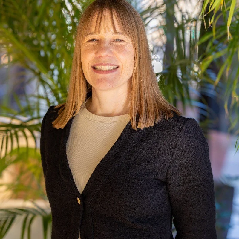 Woman with medium length brown blonde hair in professional black blazer with tan shirt
