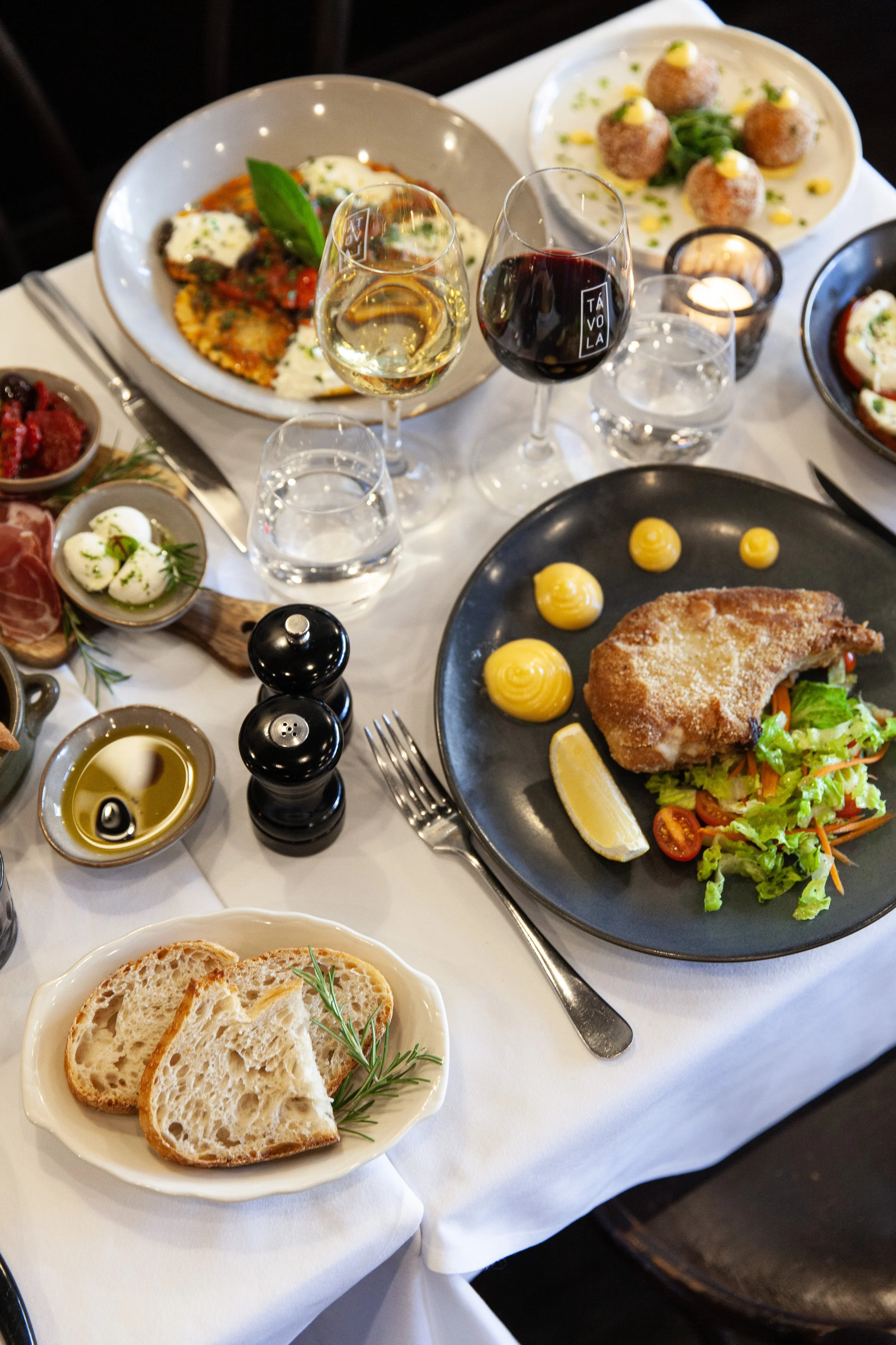 A table set with Italian dishes, including a plate with bread slices, fried chicken with salad and lemon, various small appetizers, a glass of white wine, and a glass of red wine, all arranged on a white tablecloth.
