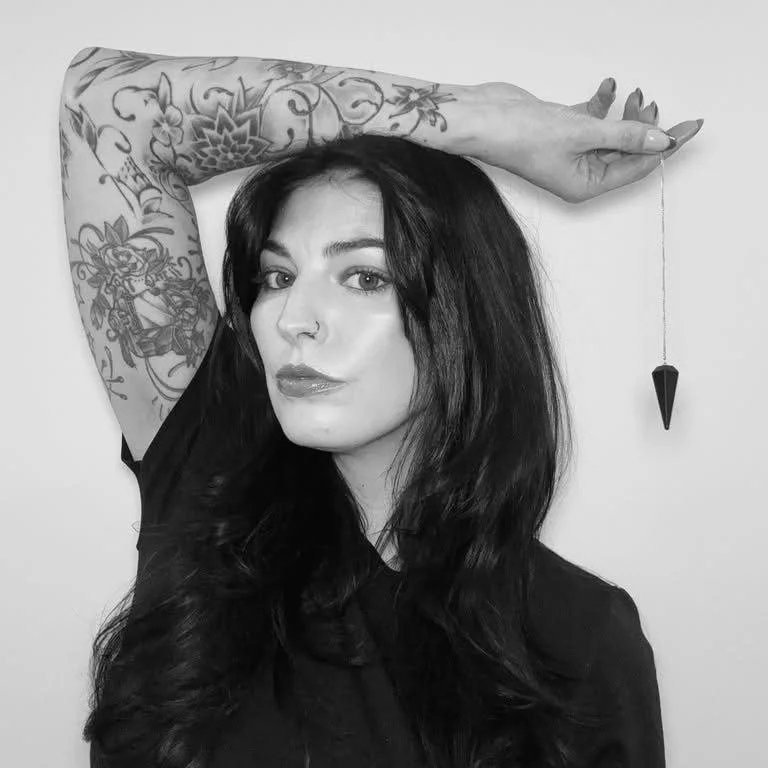 Kara Perakovic,A woman with dark hair and tattoos on her arm, posing with her arm raised above her head and holding a hanging pendulum. Host / Curator of the Oddity Shop Podcast