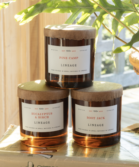 Three scented candles in amber glass jars with beige lids, labeled 'Pine Camp Ligneage,' 'Eucalyptus & Birch Ligneage,' and 'Boot Jack Ligneage,' placed on a wooden surface near a window with green leaves in the background.