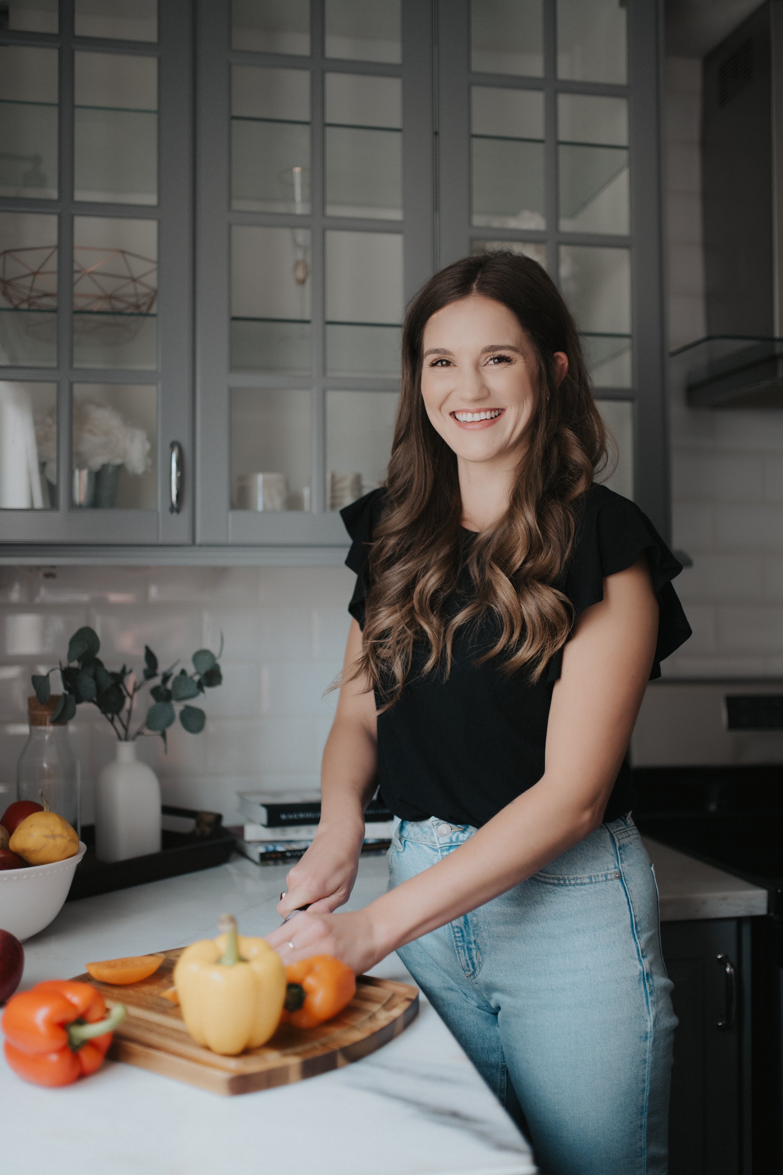 A woman with long wavy hair, wearing a black top and light blue jeans, standing in a modern kitchen, cutting yellow and orange bell peppers on a wooden cutting board, smiling at the camera.