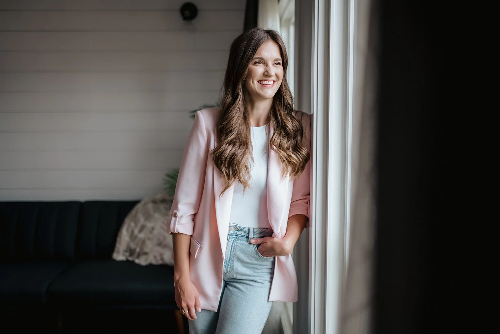 A young woman with long wavy brown hair smiling, wearing a pink blazer over a white top and light blue jeans, standing near a window with sunlight coming through.
