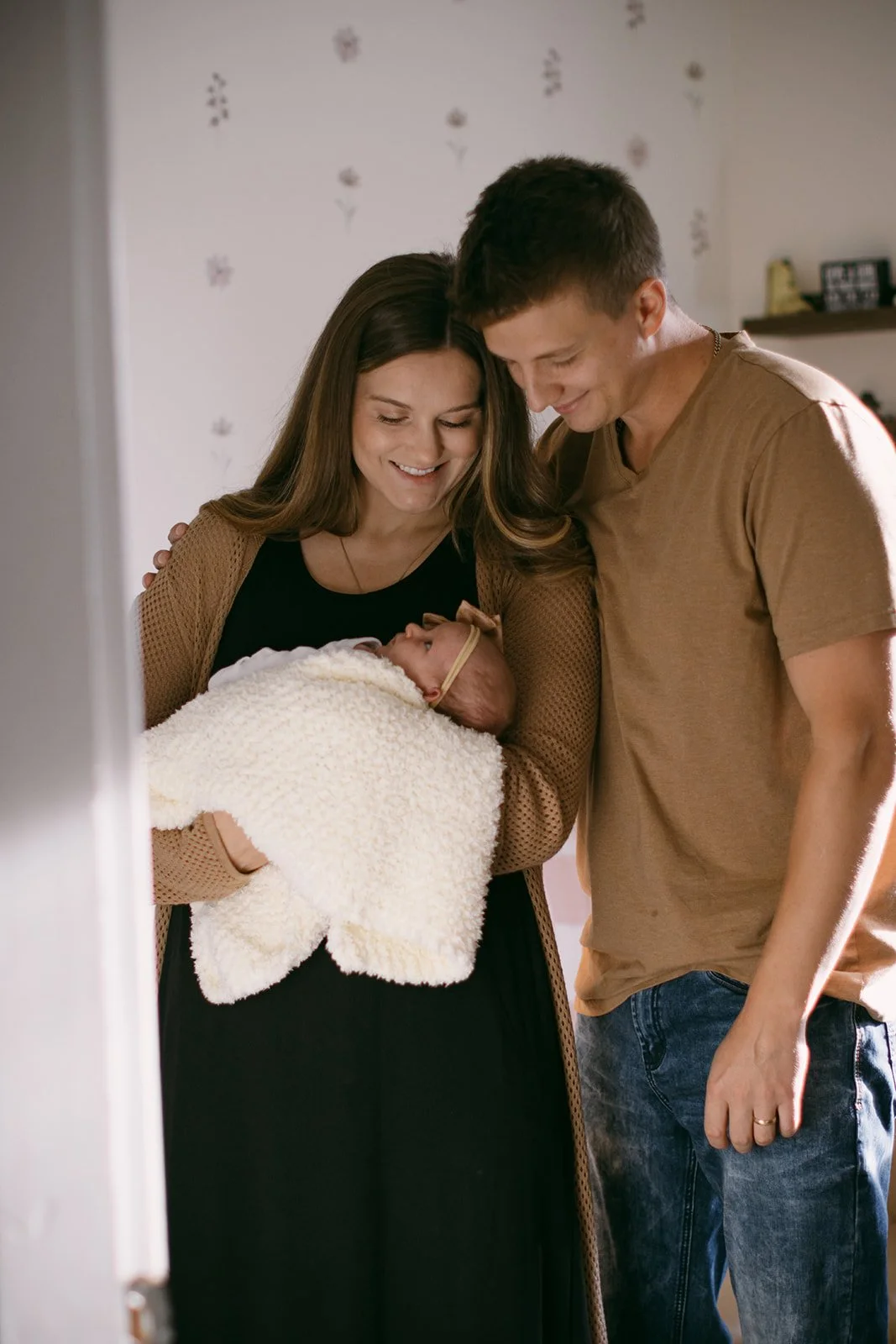A woman holding a newborn baby wrapped in a white blanket, with a man beside her, all smiling and looking at the baby in a warmly lit room.