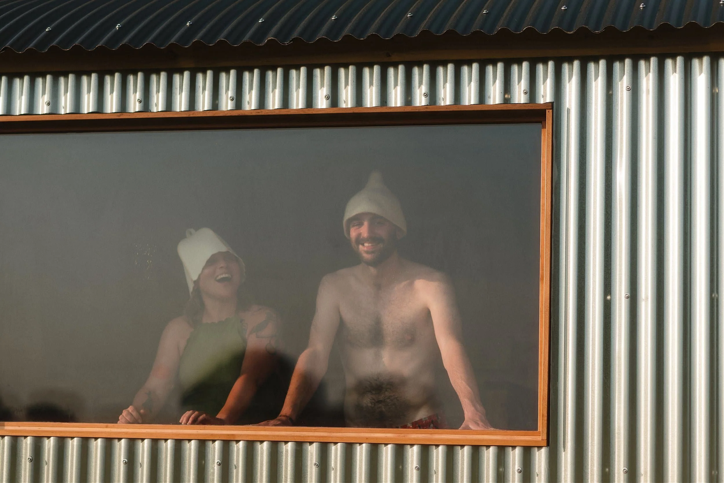 Two people, a man and a woman, are standing behind a window, both wearing Christmas hats and smiling or laughing.