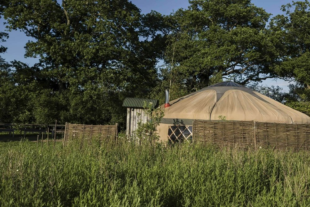 A yurt with a beige cover, surrounded by a wooden fence, is set in a grassy area with trees in the background.
