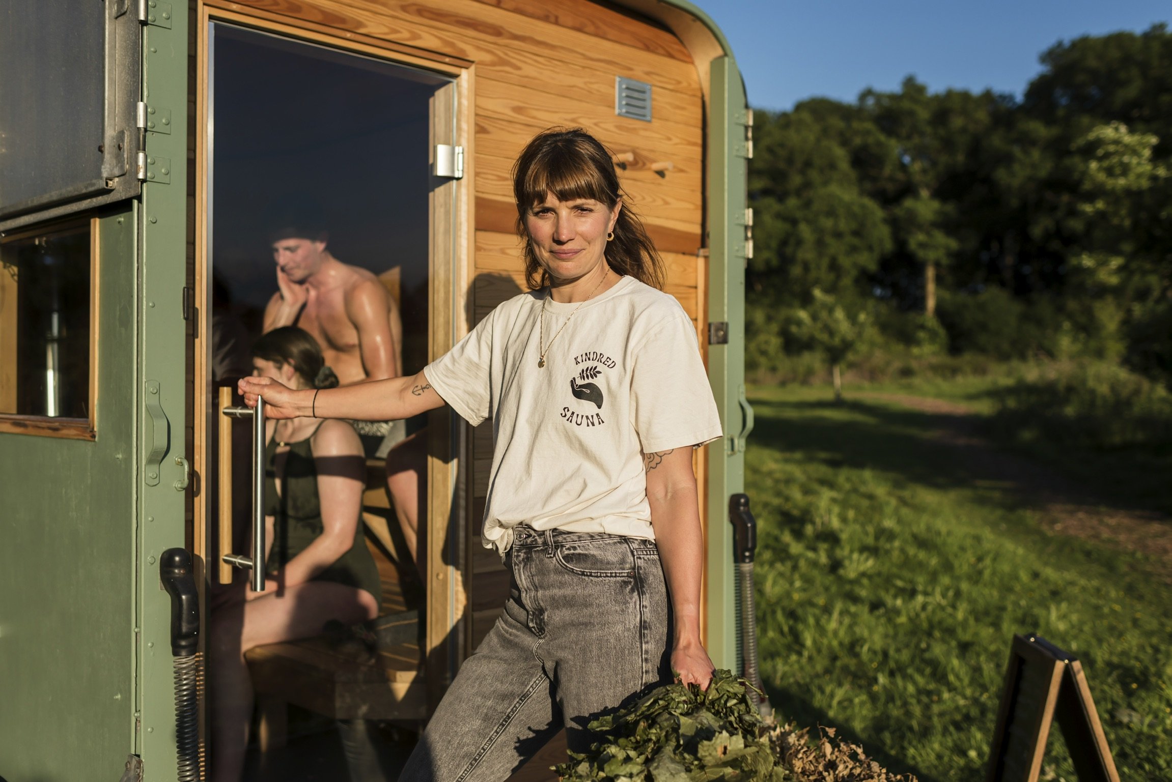 A young woman with brown hair standing outside a wooden sauna, holding a bundle of green leaves, smiling at the camera. The sauna is open, revealing a shirtless man and a woman inside, seated and relaxing. The scene is set in a green, wooded outdoor area during daytime.