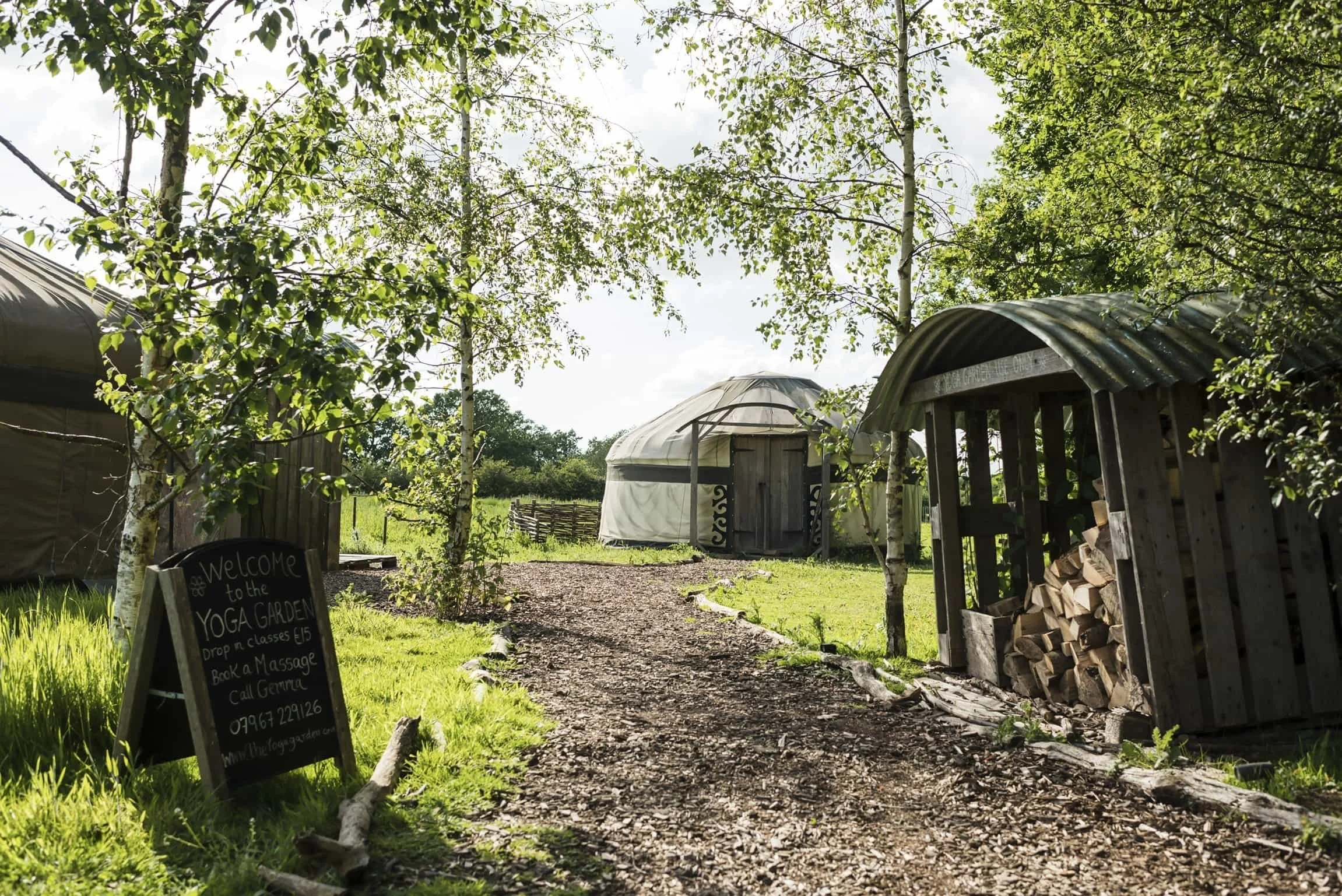 A gravel path lined with green grass and trees leads to a yurt in a rural setting. There is a blackboard sign advertising yoga classes and massages, and a wooden firewood storage with logs on the right. The sky is partly cloudy with sunlight shining 