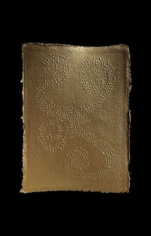 A golden-colored rectangular piece of chocolate with embossed swirls and two numbers, 2 and 3, created with small raised dots.