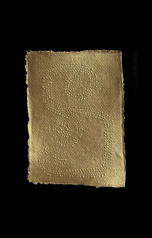 A rectangular, textured gold foil with embossed patterns of small dots forming various shapes and designs, set against a black background.