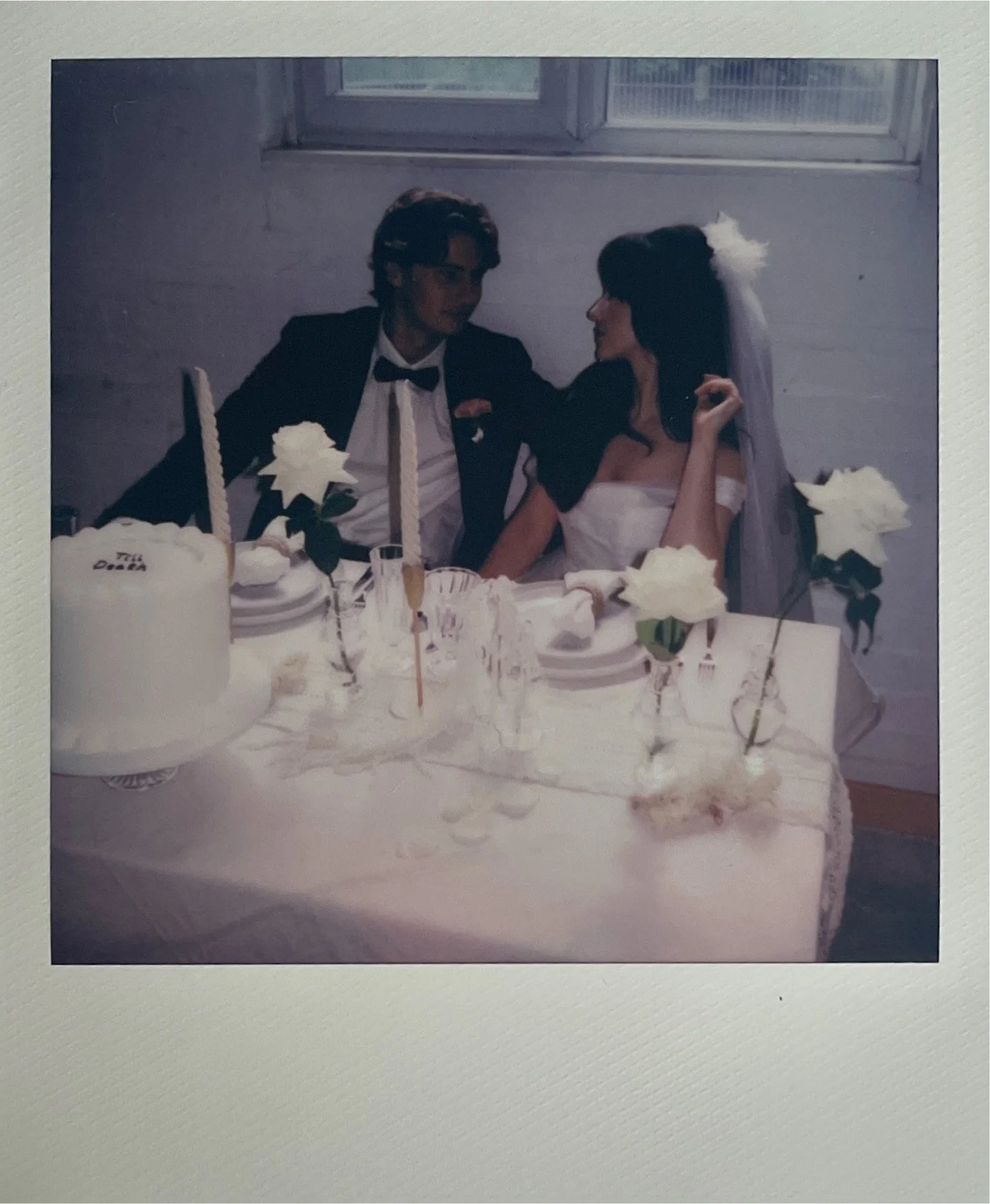 A groom and bride sitting at a wedding reception table, dressed in wedding attire, with the groom in a tuxedo and bow tie, and the bride in a white wedding dress and veil, surrounded by wedding decorations, cake, and flowers.