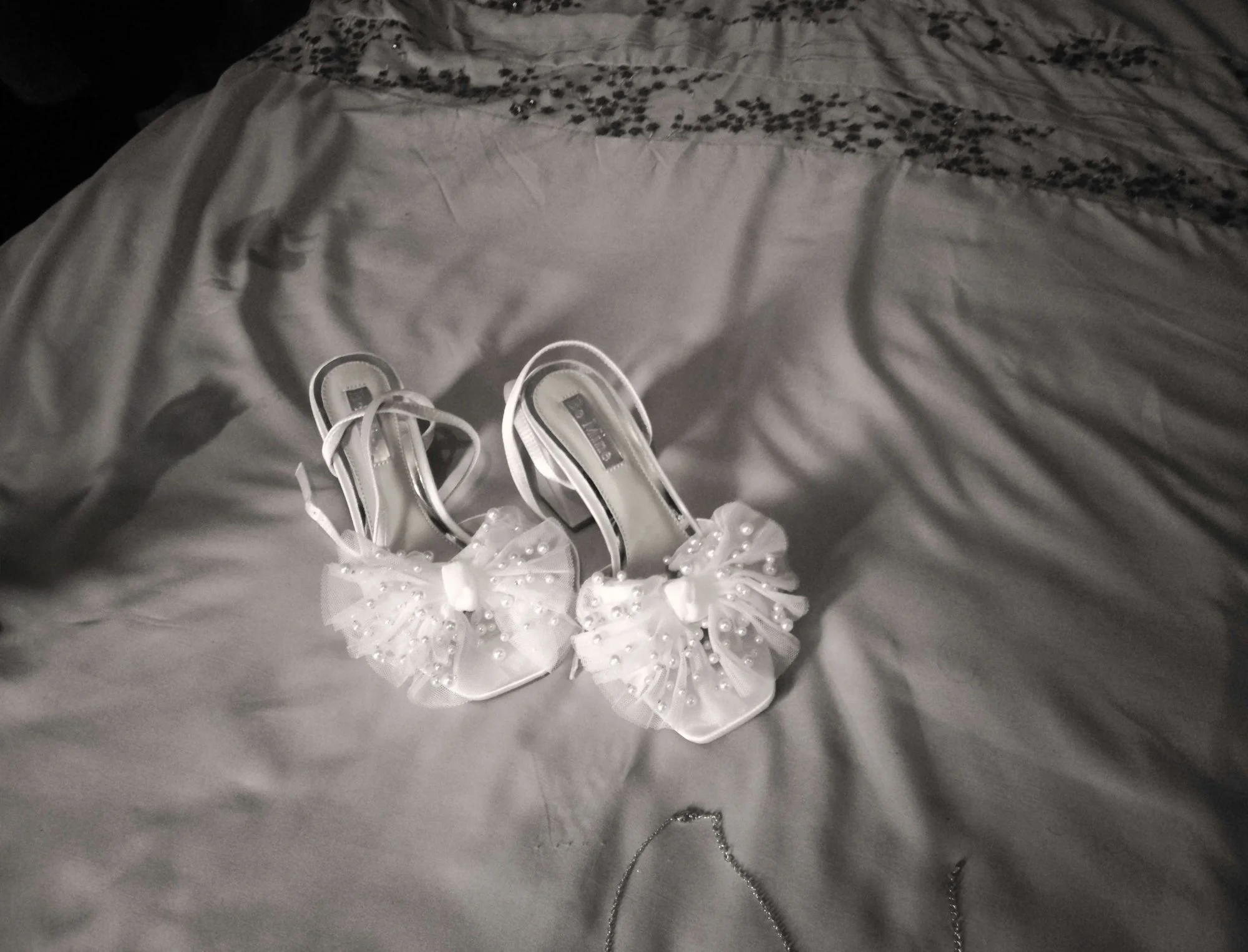 A pair of white high-heeled shoes with decorative bows and pearl accents, placed on a bed with a light-colored, slightly wrinkled sheet.